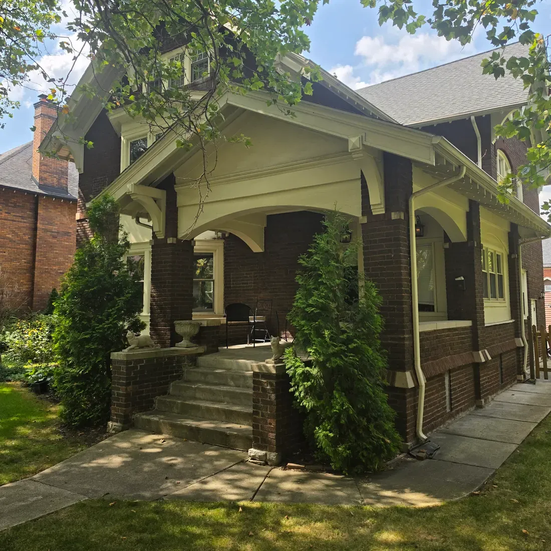 A two storey house in dark brown brick with cream painted trim. A deep covered porch with arched openings sits above concrete steps, with clipped evergreens on either side and tree canopy overhead.